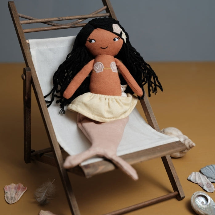 Fabelab Plush Black Mermaid doll with brown skin and black hair on a wooden deck chair with seashells