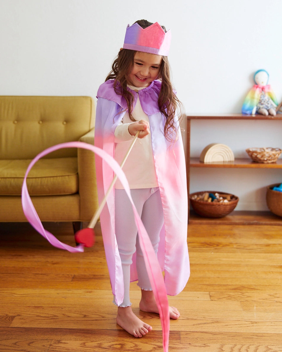 A little girl playing dress up in Sarah's Silks blossom crown, pink and purple cape, and twirling a matching heart streamer wand.