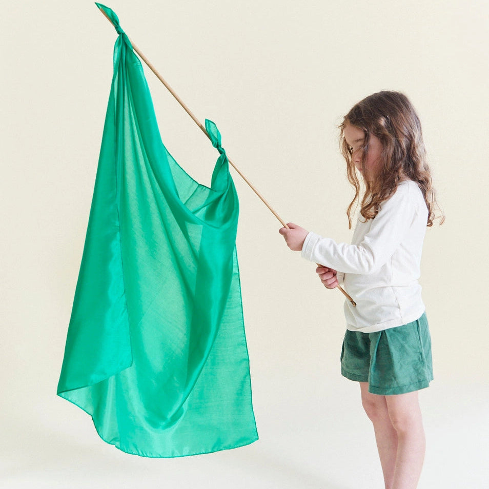 a girl holding a wooden stick with a Sarah's Silks emerald green playsilk tied to it.