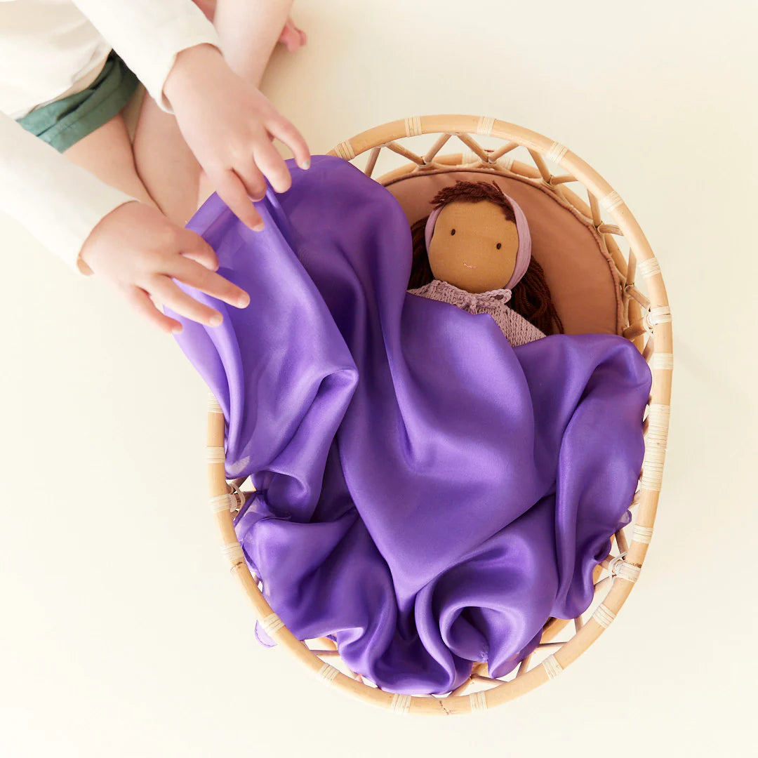 Child covering a baby doll with a solid purple playsilk