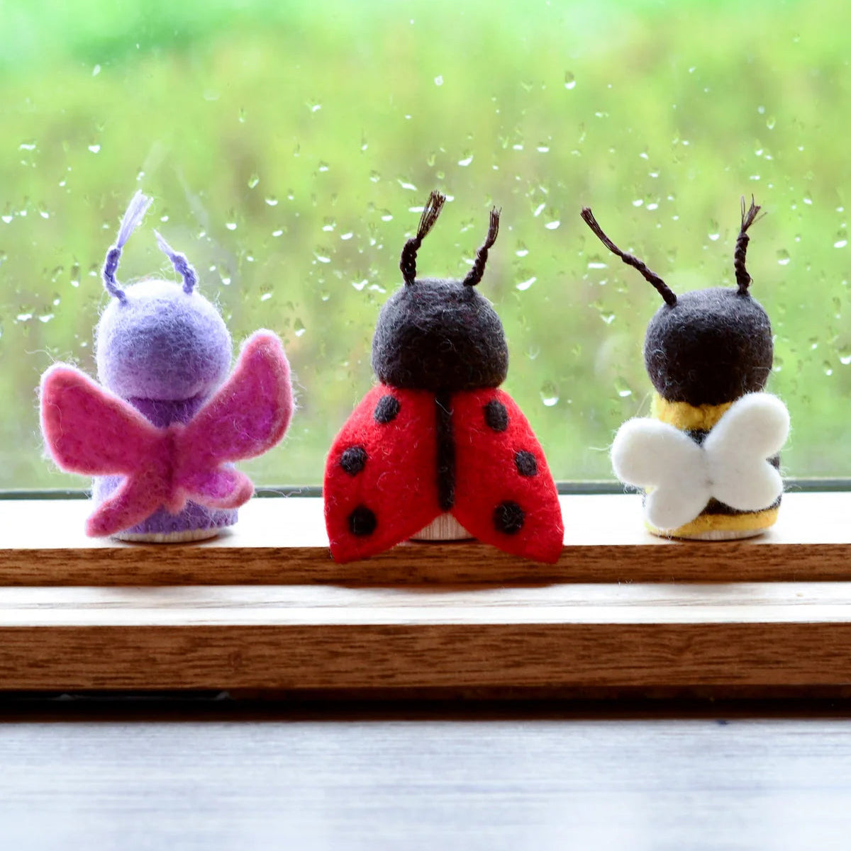 Tara Treasures felt and wood insect peg doll toys (butterfly, ladybug, bee) on a windowsill with a rainy background.