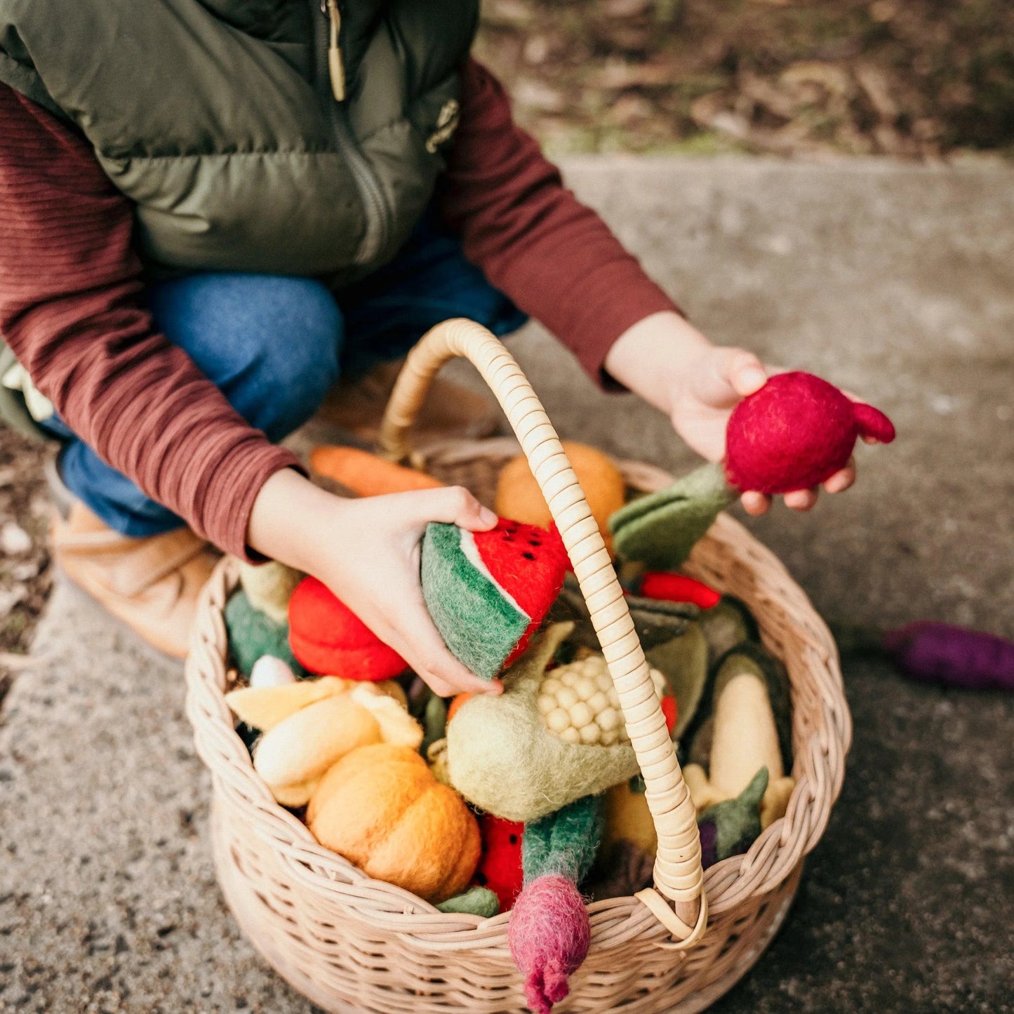 Boy holding a felt watermelon toy next to a basket of play food fruits and vegetables