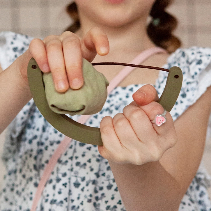 young girl holding a babai wooden toy slingshot and cotton ball in green