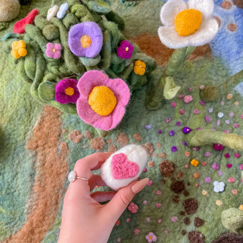 Hand holding a needle felted small  flower against a larger felted landscape with various flowers.
