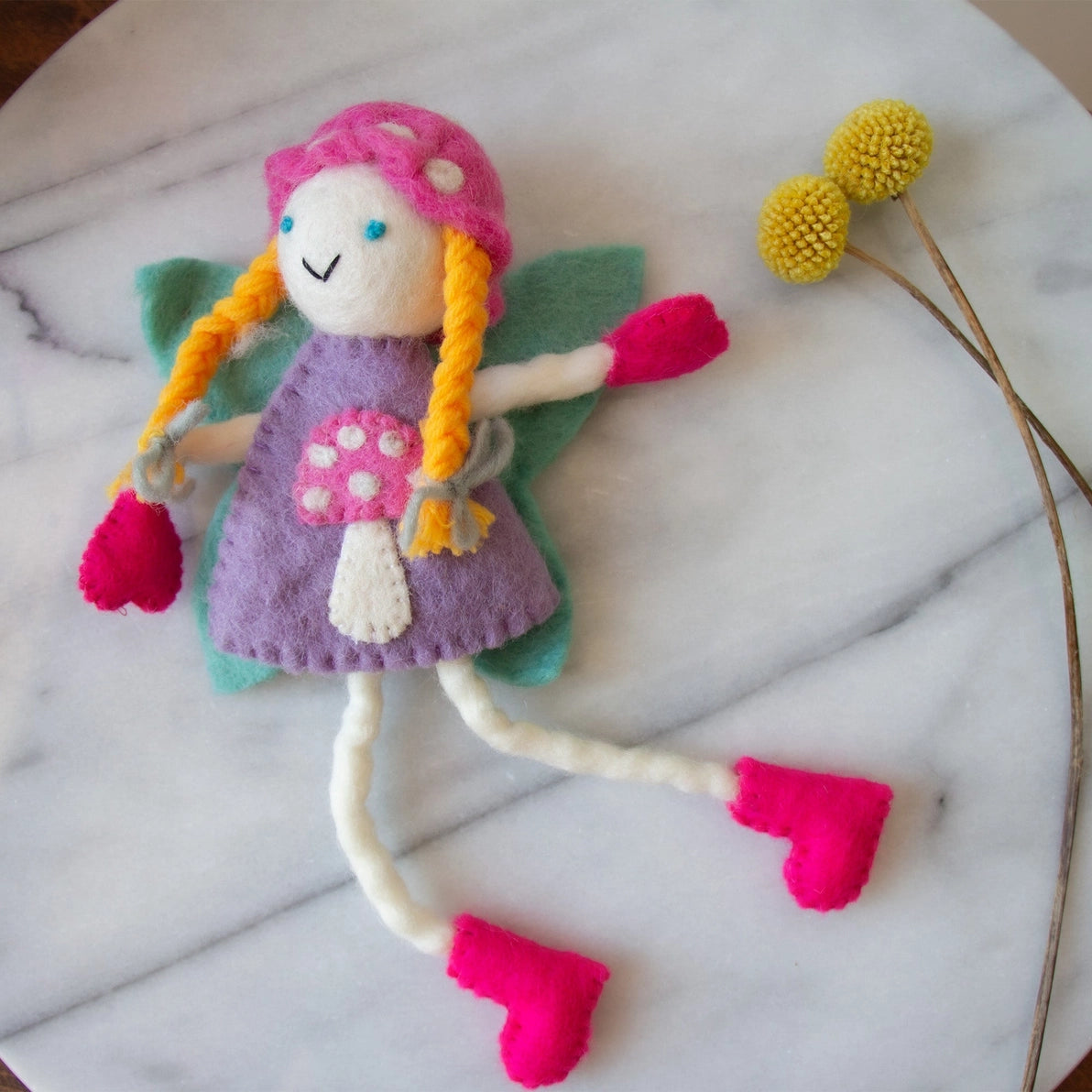Felt tooth fairy doll with blonde hair in a purple dress and pink shoes next to a flower.