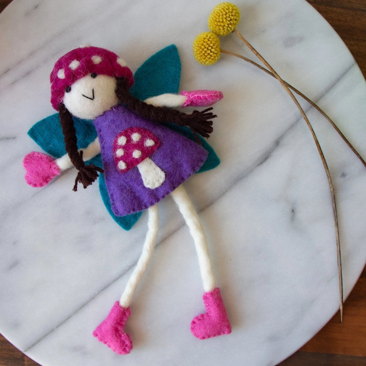 Felt tooth fairy doll with a purple mushroom dress and pink shoes next to flowers