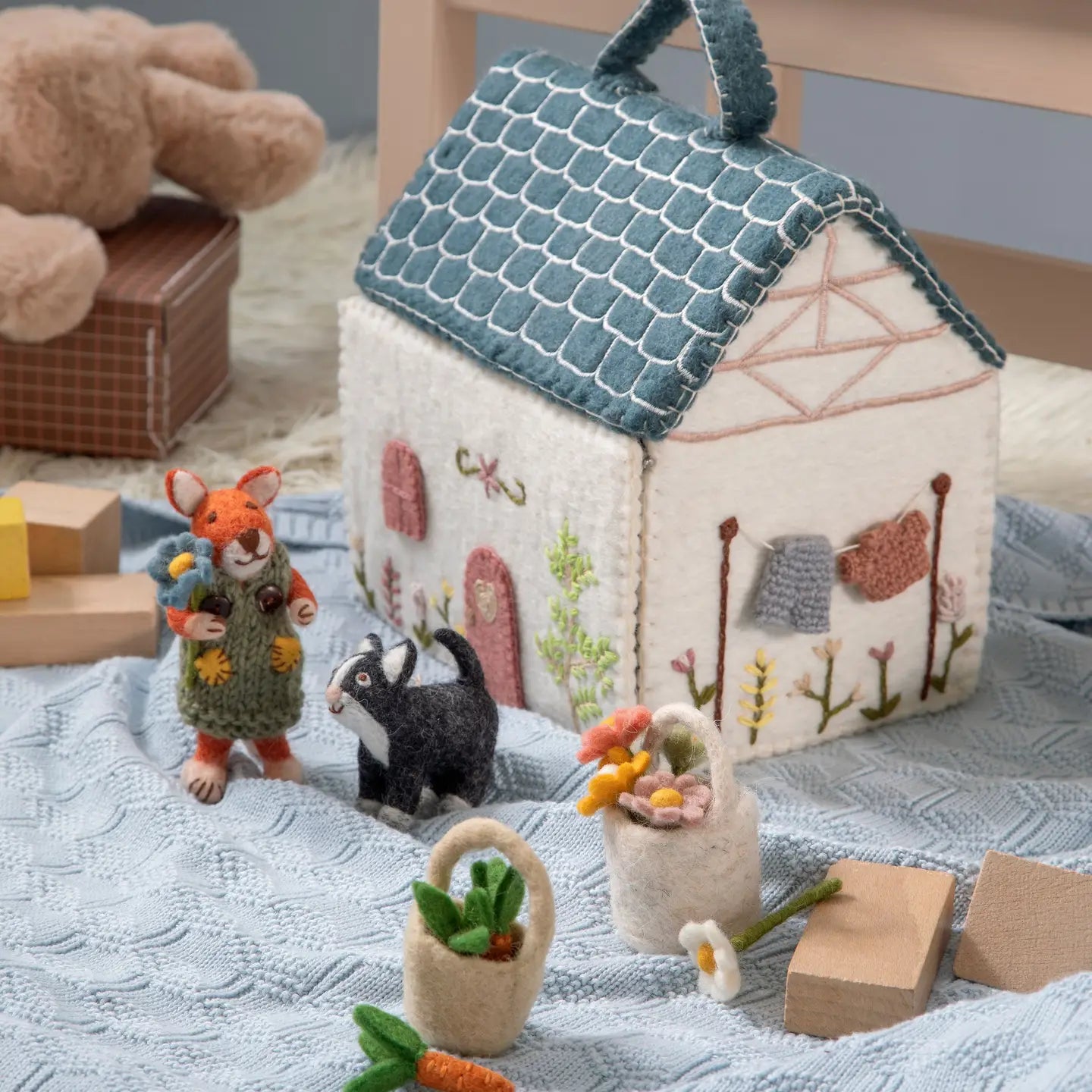 A handmade felt dollhouse with embroidered details, styled with miniature felt animals, flower pots, and toy wooden blocks.