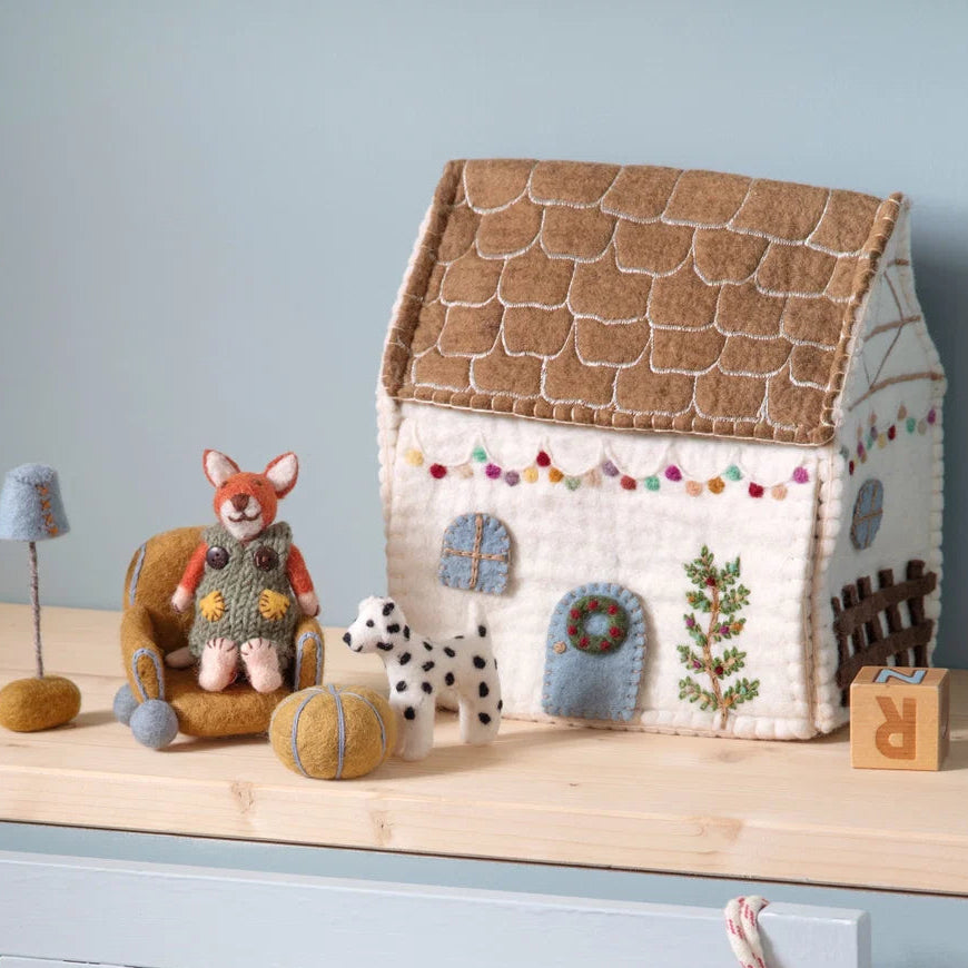 A handmade felt toy Christmas cottage dollhouse with a brown shingled roof. string lights, an embroidered Christmas tree and a wreath w alongside small felt animal figures and furniture.