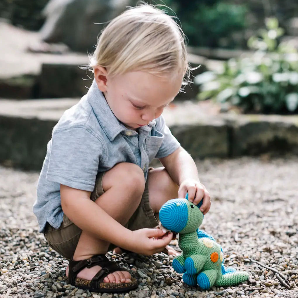 Toddler playing with a green and blue crochet toy dinosaur rattle outdoors.