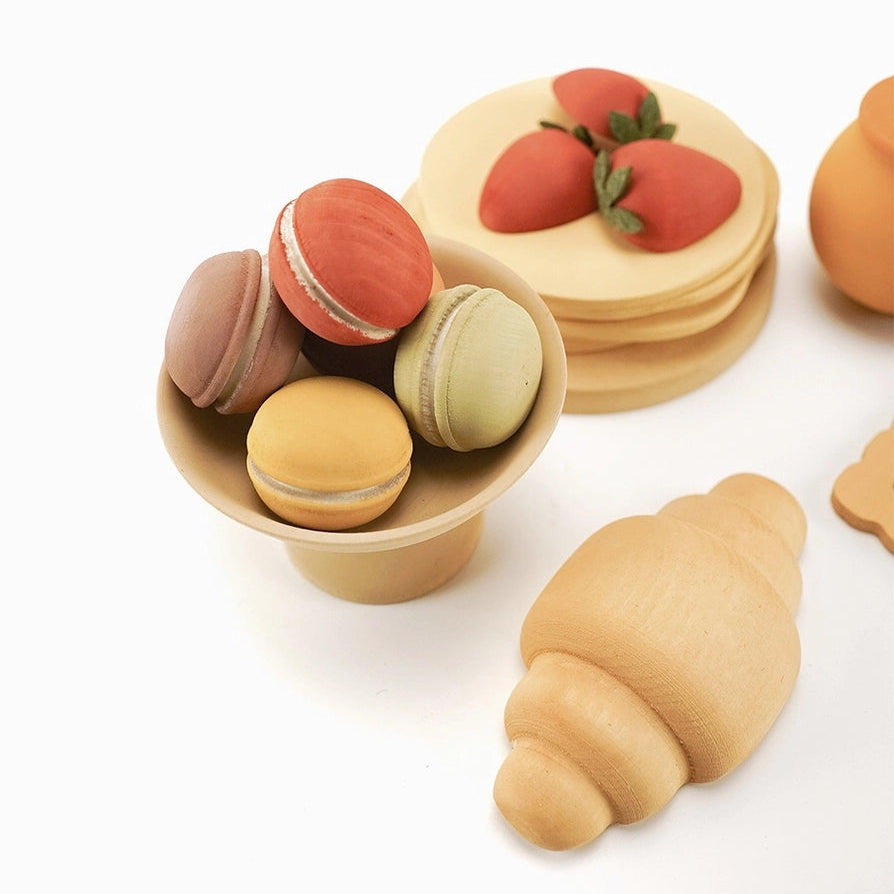 Sabo Concept wooden play food dessert pieces including macarons in a small bowl, a stack of pancakes with strawberries, and a croissant, all in a light natural wood finish