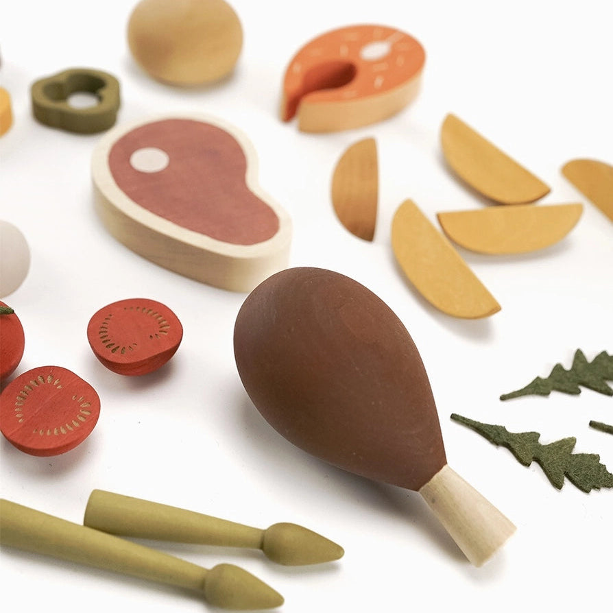 A set of wooden play food pieces from Sabo Concept including a steak, a chicken leg , sliced tomatoes, herbs, and asparagus. 