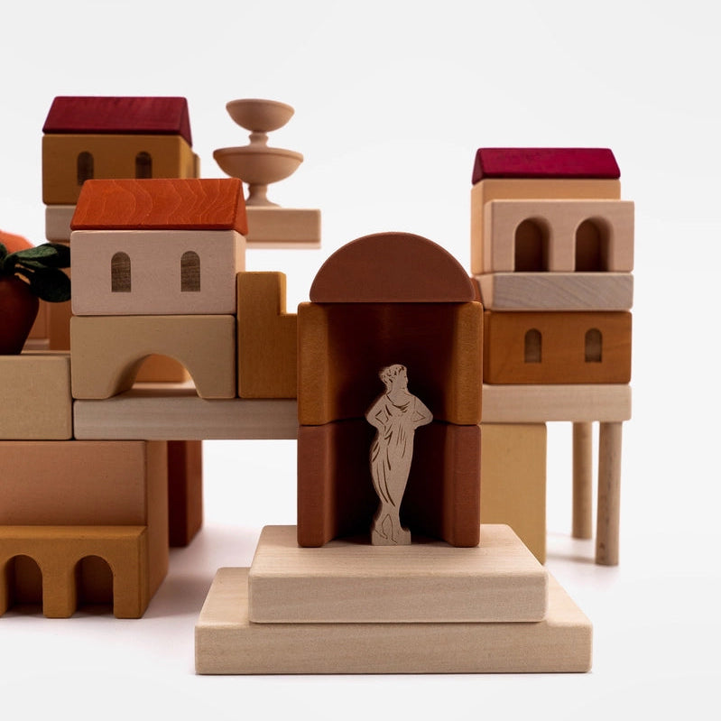 A set of wooden toy building blocks arranged to form an Italian courtyard with a Dancer statue, houses with red, orange, and burgundy roofs, arches, columns.