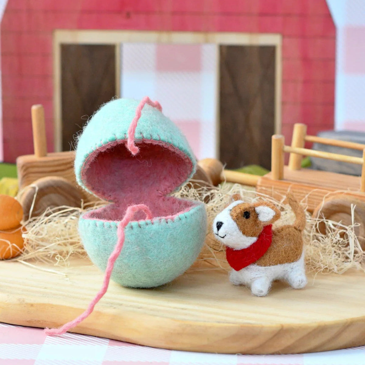 Small felt Corgi dog toy with a red scarf next to a felted Easter egg in front of a barn.