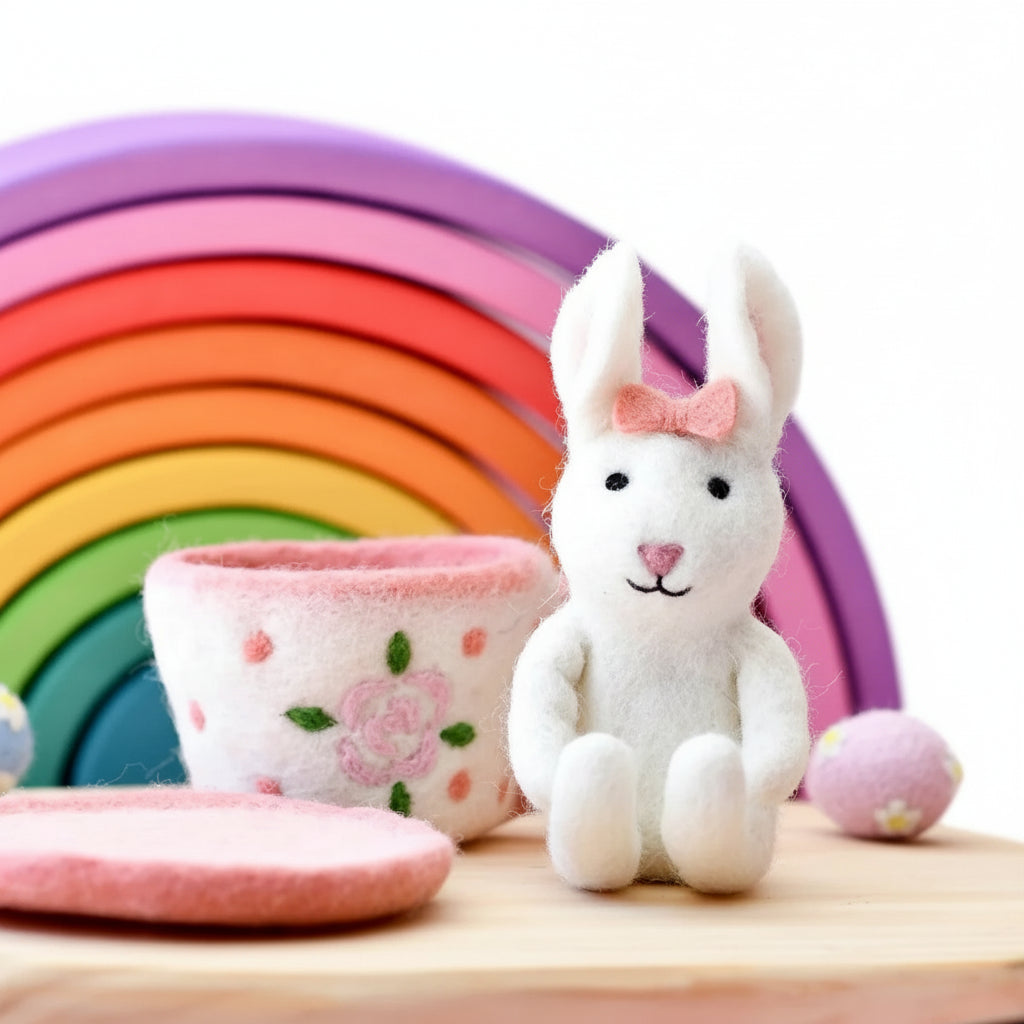 White bunny with a pink bow, colorful rainbow, and pink bowl on a wooden surface with a light purple background.