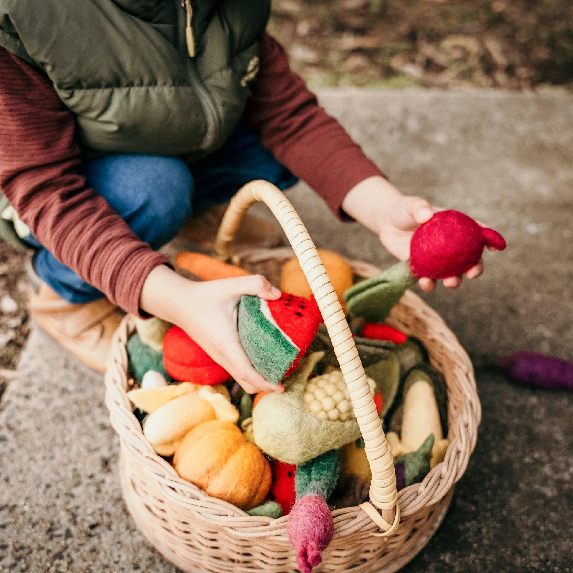 Boy holding a felt watermelon toy next to a basket of play food fruits and vegetables